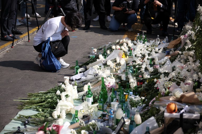A young woman in a white t-shirt kneels in front of a makeshift altar outside the Itaewon station after the Halloween crush.