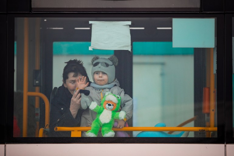 Boy is wearing hat with bear ears and holding green stuffed animal as he waves from the bus window, with his aunt helping to hold him up.