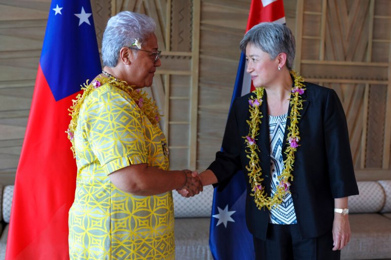 Australian Foreign Minister Penny Wong wearing a lei around her neck and in a black suit shakes hands with Samoa's Prime Minister Fiame Naomi Mata’afa who is wearing a yellow dress. There are flags in the background.