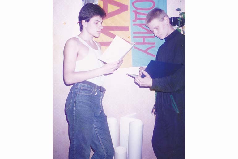 A photo of Sergej at university in a tank top and jeans, reading a piece of paper/book, with a person standing in front of them.
