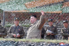 Kim Jong Un in a sandy coloured military jacket gesturing with his left hand and looking serious with senior military commanders at an outdoor location