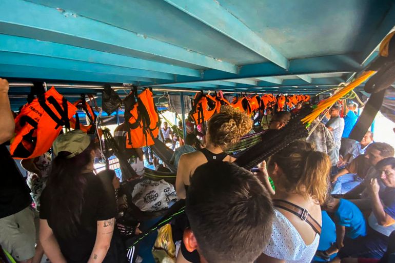 Tourists crammed into a low-ceilinged boat, some standing some sitting, while orange life jackets hang along the side