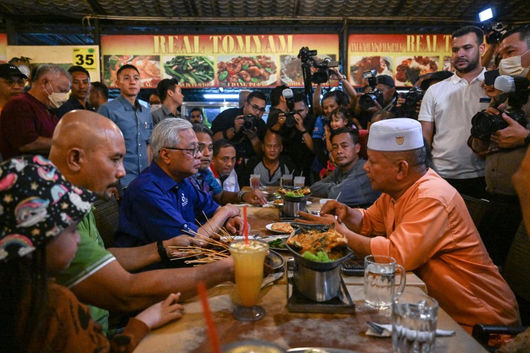 Malaysian Prime Minister Ismail Sabri Yaakob sitting at a long table opposite a voter and surrounded by media during a campaign stop at a restaurant.