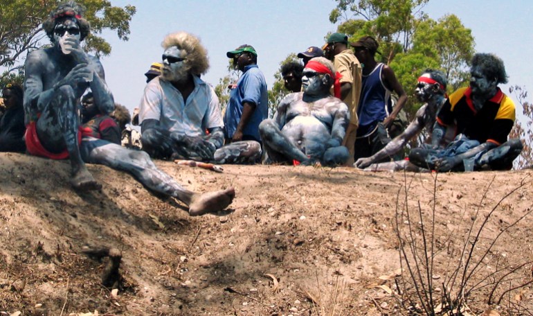 Indigenous men, some with body paint, sitting down and listening to speeches on sandy ground in the Northern Territory of Australia