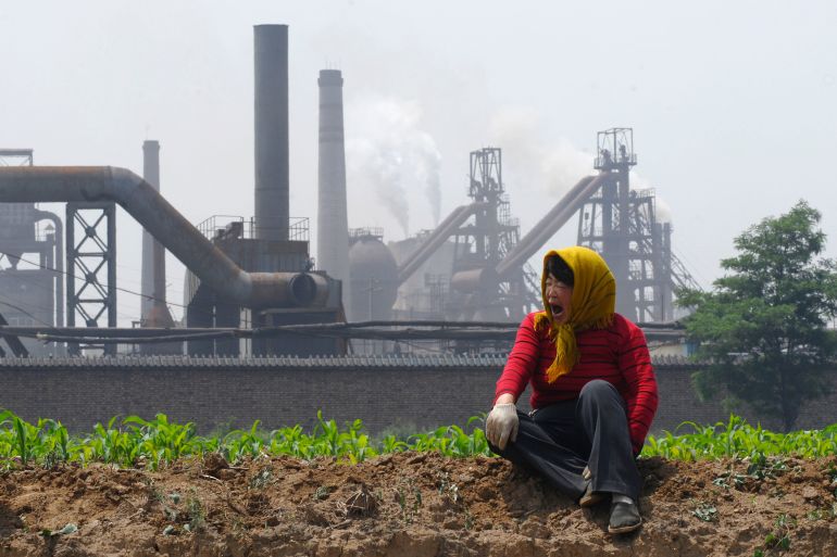 Chinese woman sitting on the side of the road, yawning, wearing yellow headscarf, with a factory in the background releasing fumes