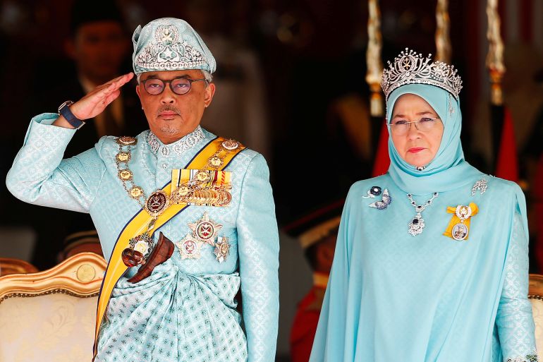 Malaysia's king and queen in pale blue traditional outfits. The king is saluting.