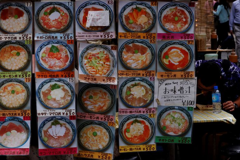 Window of restaurant in Japan displaying menu: photos of dishes with prices. A man can be seen eating noodles inside