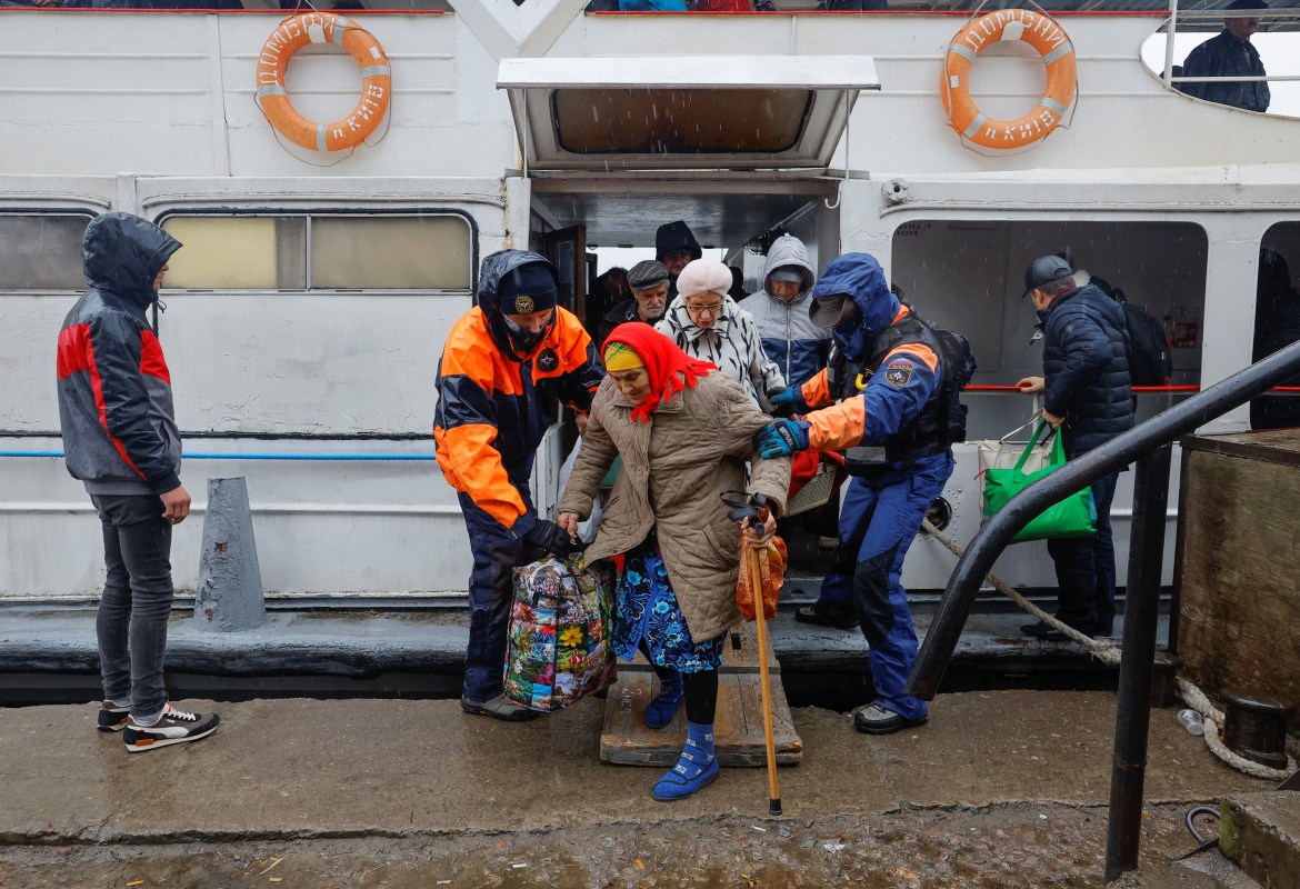 Elderly women, one wearing a red headscarf and using a walking stick while holding bag, being helped to come off ferry