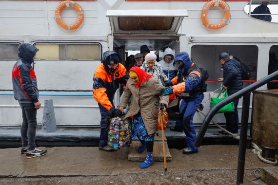 Elderly women, one wearing a red headscarf and using a walking stick while holding bag, being helped to come off ferry