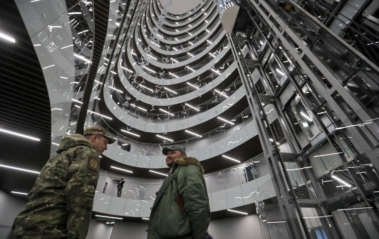 Camera looks up at two men in military uniform stand inside building and its many floors, curving around them
