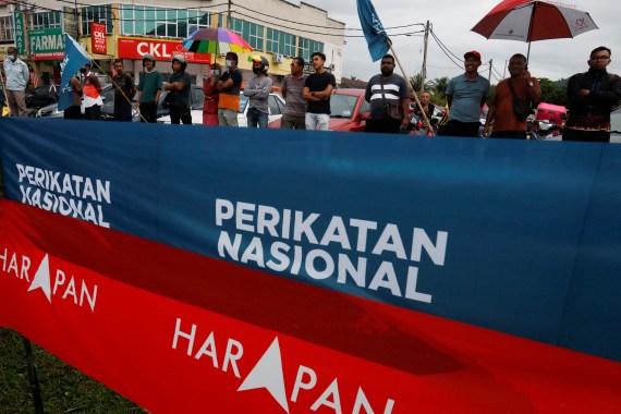People standing behind a red banner for Pakatan and teal blue banner for Perikatan as they watch a political rally in a Malaysian town.