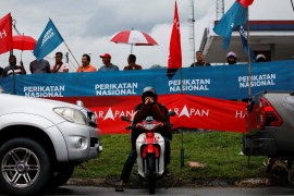 A man on a motorbike with banners for rival parties behind him and other people watching