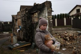 Girl looking sad, wearing a winter jacket and hat, holds a ginger cat. Behind her is one remaining wall of a destroyed house with rubble and bricks surrounding it.