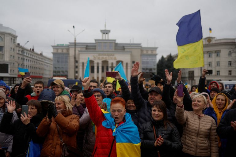 A crowd gathers in the Kherson city centre, some wrapped in Ukrainian flags or flying them, looking happy.