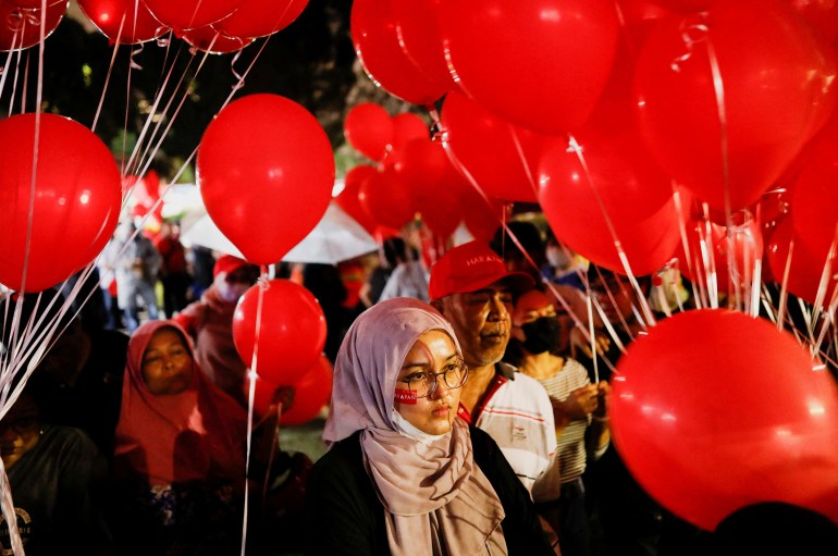 Een jonge vrouw tuurt aandachtig tussen een menigte mensen met rode ballonnen tijdens een politieke bijeenkomst voor Pakatan Harapan