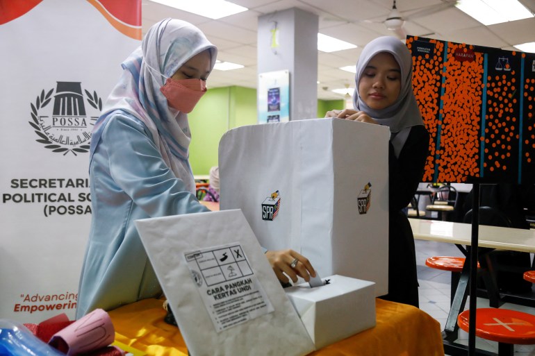 Een jonge student met hoofddoek en masker legt uit hoe het Maleisische stemsysteem werkt in een nagemaakt stembureau op een universiteit