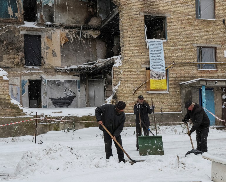 Local residents shovel snow as a work of world-renowned graffiti artist Banksy is displayed on the wall (L) of a destroyed building in the Ukrainian village of Horenka, which was heavily damaged by fighting in the early days of the Russian invasion, November 19, 2022.