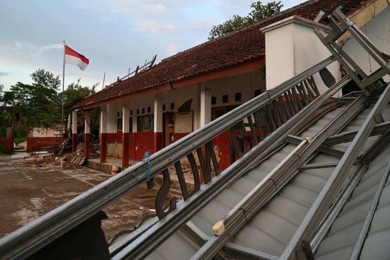 A damaged school building after the earthquake with a metal structure collapsed onto the roof and rubble lying about the main school building while an Indonesian flag is still flying.