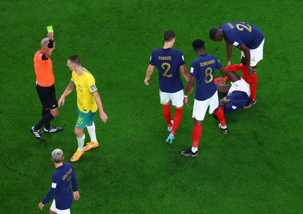 From the top, ref holds up a card as Duke walks past him. Behind him three French players crowd over Dembélé on the ground