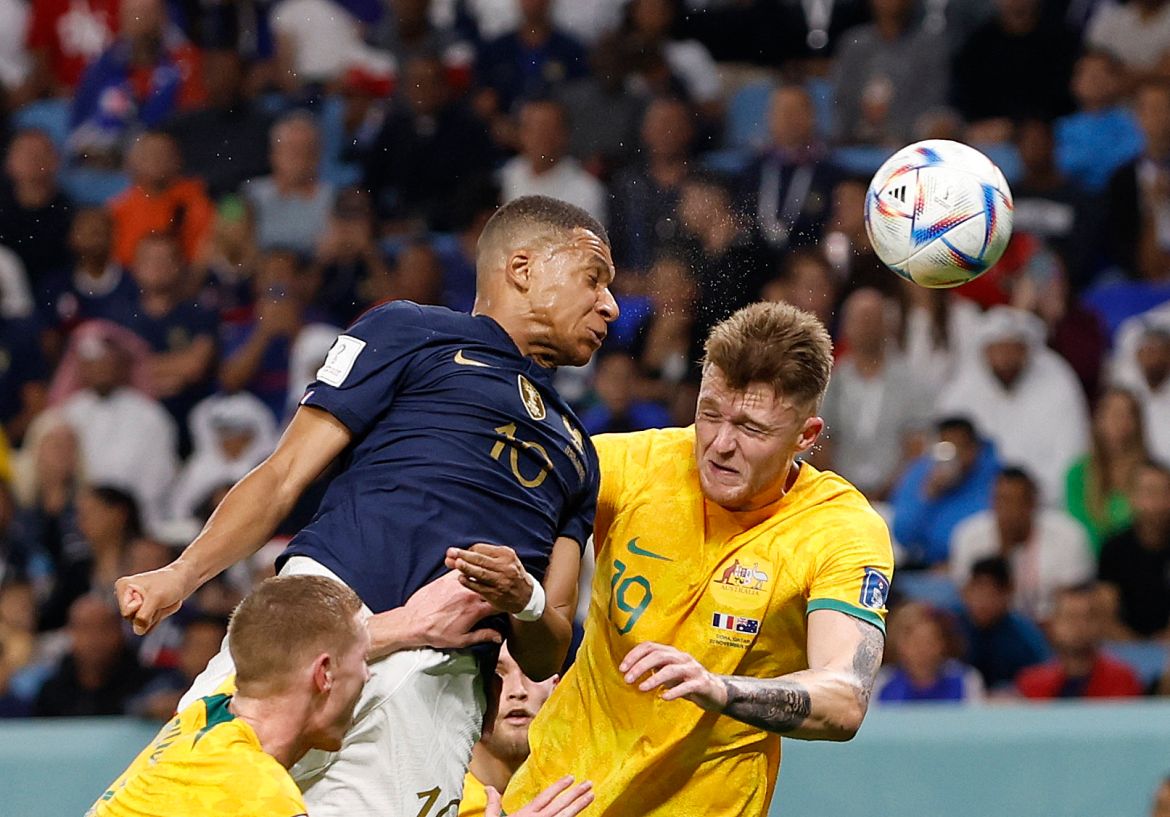Mbappe in mid-air, face contorted after having hit the ball with his head, sweat flying around them, and two Australian players on either side of him with equally contorted faces