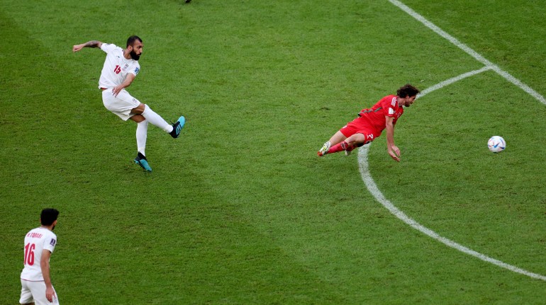 Iran's Roozbeh Cheshmi in action, just kicking the kick, as the Welsh defender looks behind him as the ball has flown past him on its way to goal