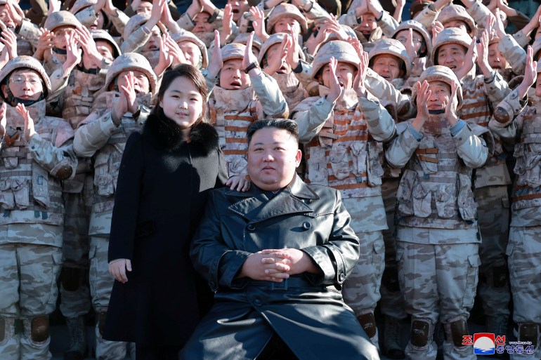 Kim está sentado olhando para a câmera enquanto sua filha, vestida com um casaco preto, está à sua direita com a mão em seu ombro.  Atrás deles, estão várias fileiras de homens vestidos com roupas militares, batendo palmas com as mãos bem para o alto - alguns acima da cabeça