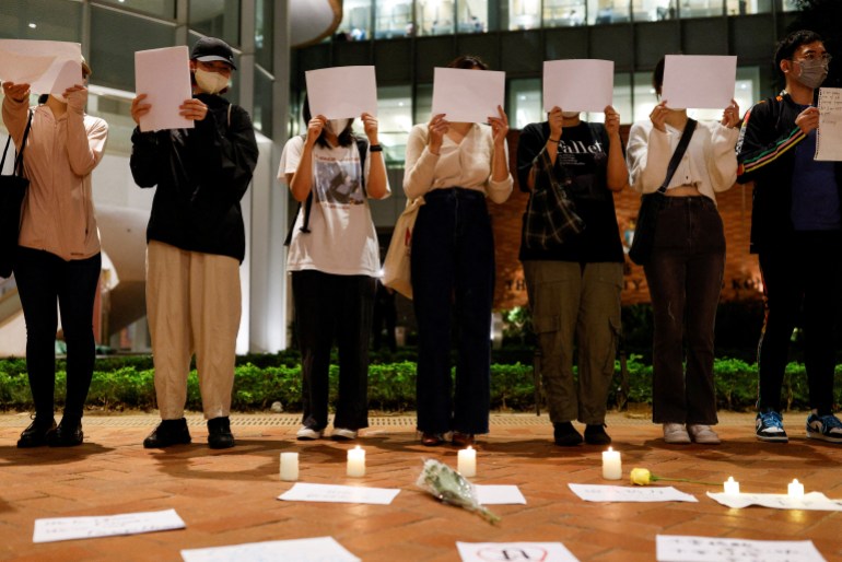 Pengunjuk rasa mahasiswa di Hong Kong memegang lembaran kertas kosong sebagai bagian dari kewaspadaan atas kebijakan COVID-19 di Tiongkok. Mereka memakai topeng dan sebagian besar wajah mereka tertutup. Ada lilin menyala di lantai di depan mereka.