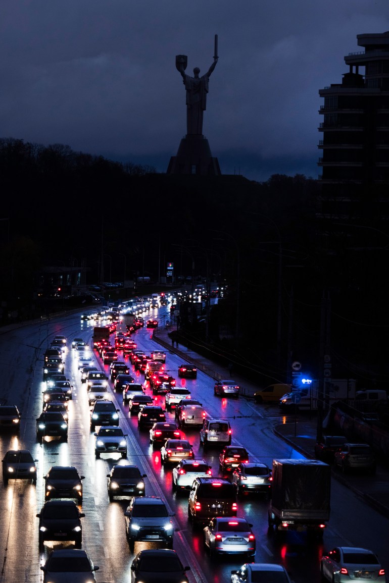a main road showing cars' white headlights and red rear lights in the darkness of Kyiv with Motherland Monument in the background