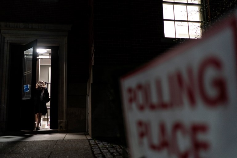 Woman walking into door of building which casts a light onto the path outside and the close-up blurred sign saying: 'Polling place'