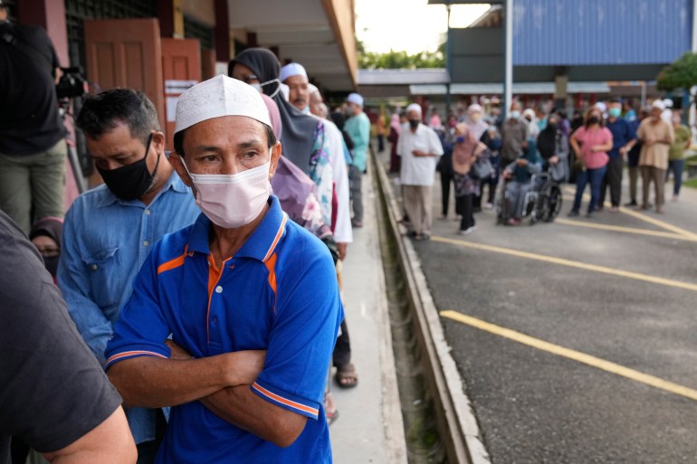 A man wearing a blue t-shirt, a white skull cap and a face mask crosses his arms as he stands in front of a voting line in Malaysia.