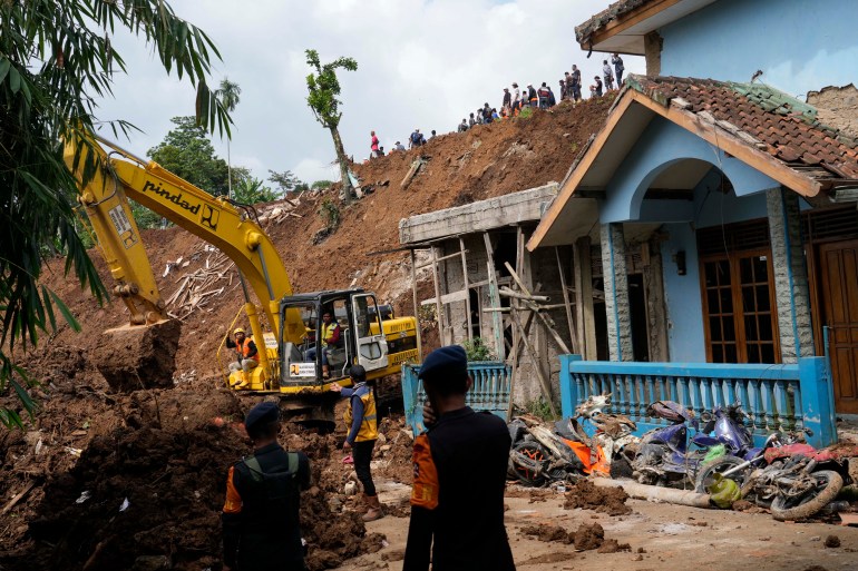 Rescue workers, looking small, stand atop a giant mound of mud from a landslide with a badly damaged house in front of it.  An excavator is also used.