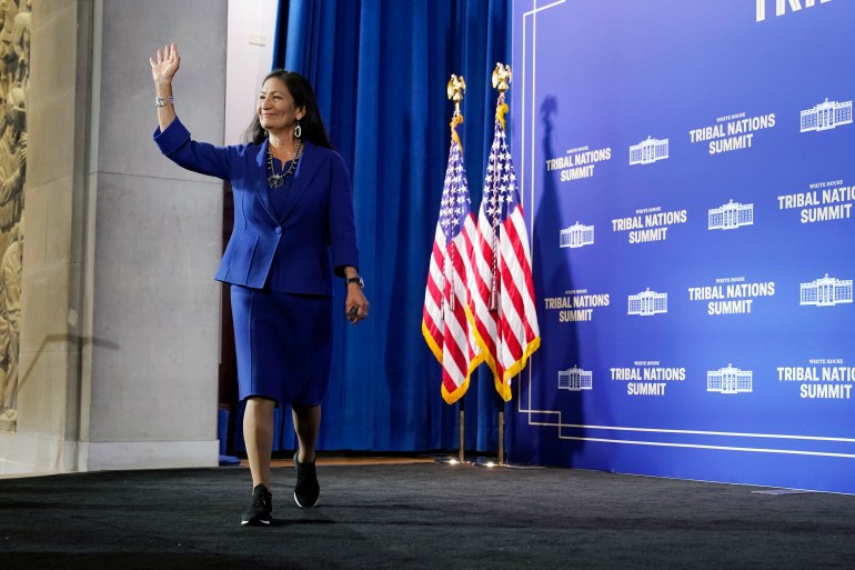 Deb Haaland waves from the stage as she moves forward, with two American flags behind her
