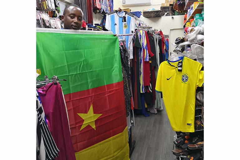 A photo of a shop with various shirts and hair extensions and a man standing in front holding a Cameroon flag.