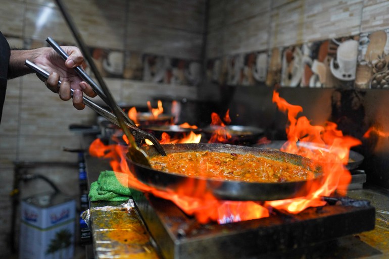 A cook holds a karahi with a plier-like implement over a roaring flame
