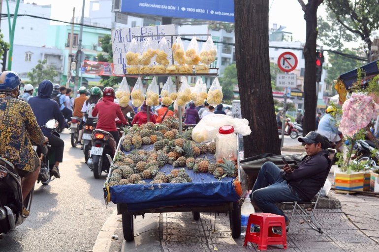 A fruit vendor by the roadside in Ho Cho Minh City. Whole pineapples are laid out on the cart with cut pineapples hanging from plastic bags in front. He's looking at his phone as he waits for customers