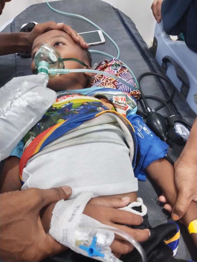 Mohammed Fajar lying on a hospital bed with a canula in his hand and breathing apparatus over his face as a family member comforts him
