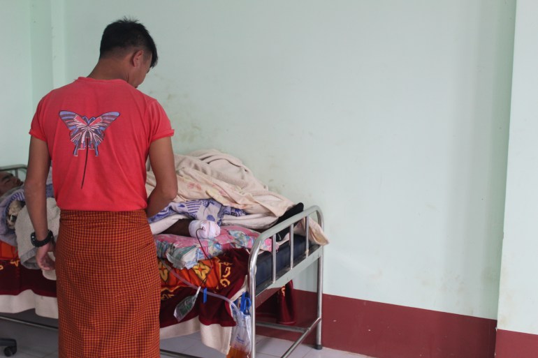 A man in red t-shirt and checked longyi looks at the bandaged leg of a fighter after an amputation. The fighter is lying in a metal-framed bed beneath mounds of blankets
