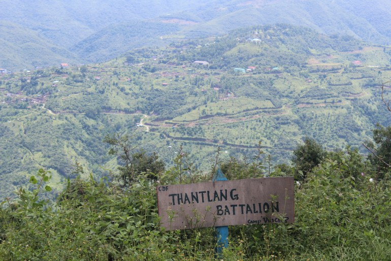 A wooden sign with Thantlang Battalion written on it in black and a view of mountain sides and some buildings in the distance