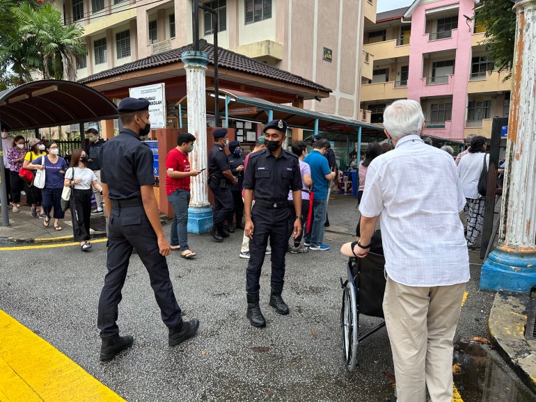 Un homme pousse un électeur en fauteuil roulant dans un bureau de vote à Kuala Lumpur avec des policiers debout à l'entrée et une file d'électeurs à gauche