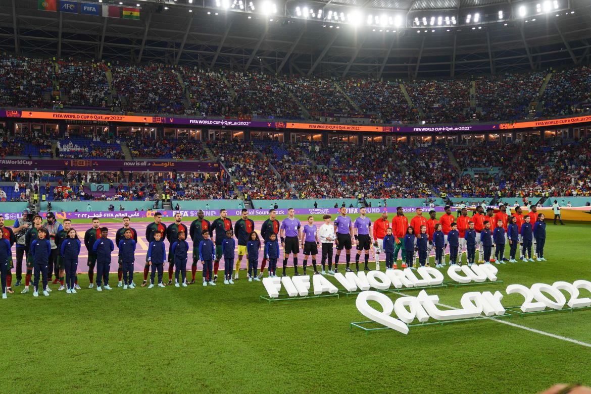Both teams in a line on the pitch with the stalls of fans behind them. In front are large white letters lying on the ground saying: "Fifa world cup Qater 2022"