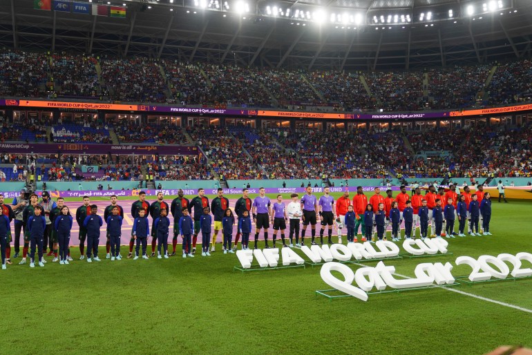 兩支球隊在球場上排成一排，球迷的座位在他們身後。 前面是躺在地上的白色大字，上面寫著： "2022年卡塔爾國際足聯世界杯"