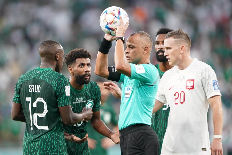 Polish and Saudi Arabian players surround a referee who is holding the ball during their match.