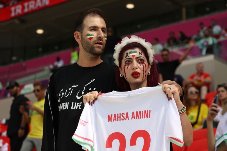 Iranian fan with face paint imitating bloody tears holds up an Iran national team jersey with Mahsa Amini 22 printed on the back. A man in a black shirt with 'women, life' printed on stands behind her