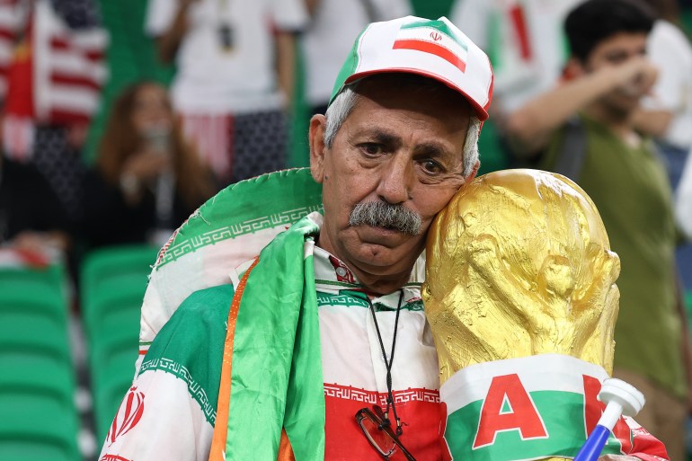 Man wearing clothes and cap with Iranian flag colours, leans his head against the trophy replica with a very sad look on his face