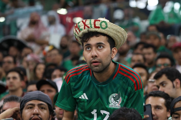 A Mexican fan wearing the team shirt and a straw hat with Mexico written in red and green on the upturned brim watches the match.  He looks tense