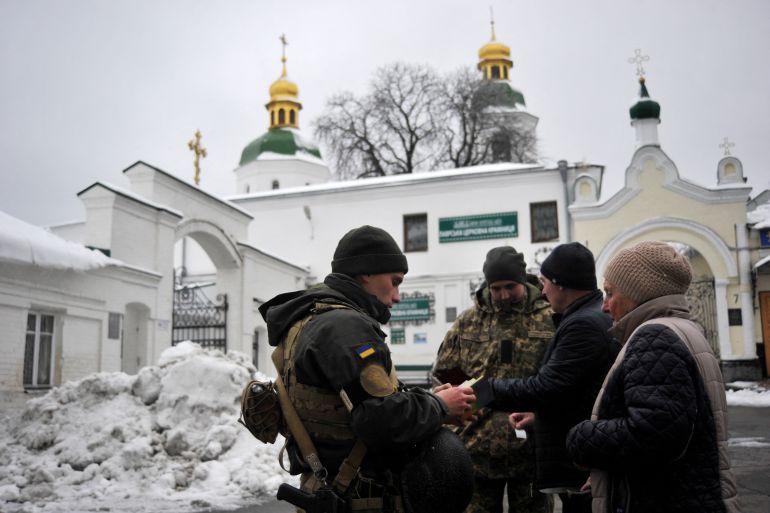 A group of intelligence officers checking documents of visitors outside the Pechersk Lavra monastery in Kyiv. Two golden domes are visible and there is snow piled up behind them. It looks cold