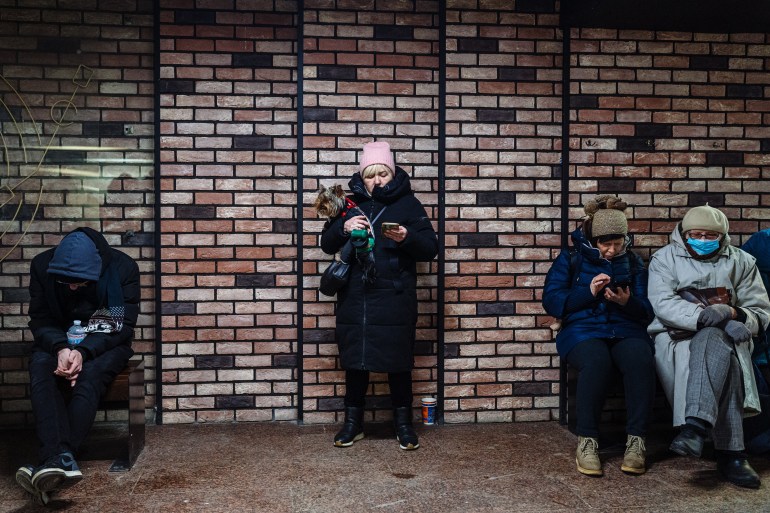 Ukrainians take shelter from Russian missile attacks in an underground passageway. They are dressed in thick coats and hats and three are seated on benches while one is standing