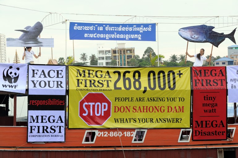 A wall of billboards and posters targeting Mega First, the Malaysian company that built the Don Sahong Dam.  The signs have big red signs like 'stop' as well as 'face your responsibilities' and 'catastrophe'.  One activist on the right is holding a large fish and the other on the left is holding a large dolphin named Mekong 
