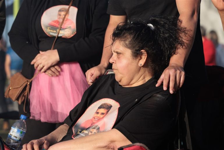 Mechelle Turvey sits in a chair wearing a black t-shirt with a white circle in the middle holding Cassius's photo. People behind her stand in the same t-shirts