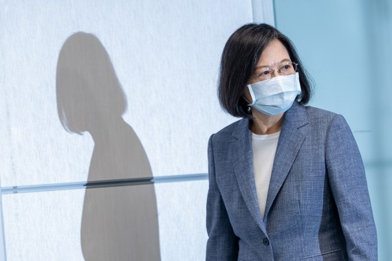 Taiwan President Tsai Ing-wen wearing a sombre grey suit. She is walking and looking to one side and casting a shadow against the wall. She is also wearing a face mask
